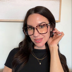 A woman with long dark hair wearing Cougar cheetah tortoise oversized square eyeglasses, a black short-sleeve top, and gold necklaces, posing indoors in front of a framed landscape print on a light wall.