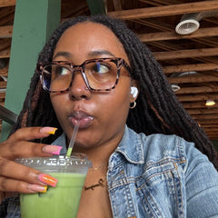 A close-up lifestyle shot of a woman with long dark dreadlocks and Cougar oversized cheetah tortoise glasses, wearing a light blue denim jacket and holding a green iced coffee in a clear plastic Starbucks cup with a clear straw.