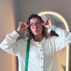 A woman posing in front of a mirror wearing a white textured cardigan and matching mini skirt, accessorized with a bright green crossbody bag and Marion blue light olive green glasses.