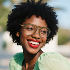 A smiling woman with curly black hair wearing Marion Aviator olive green-framed eyeglasses and a light green blouse, photographed outdoors with a softly blurred background.