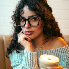A woman with curly dark hair wearing thick Amber Tortoise square eyeglasses, a light blue off-the-shoulder knit sweater, and a gold necklace, holding a takeaway coffee cup in front of a white brick wall.