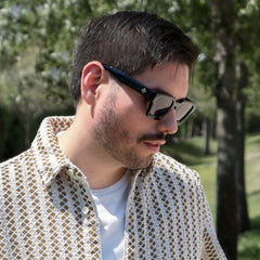 A man wearing a beige short-sleeve button-up shirt with a black geometric pattern, layered over a white t-shirt and wearing River black sunglasses, standing outdoors in a park with trees in the background.